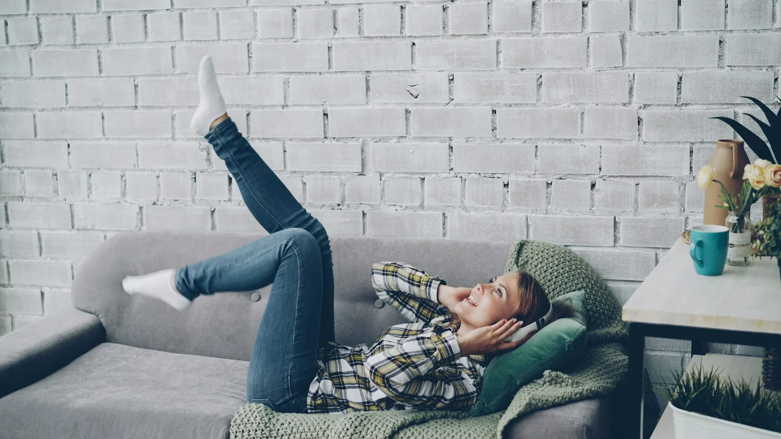 Smiling woman in headphones relaxes on a grey sofa, enjoying free time away from cleaning