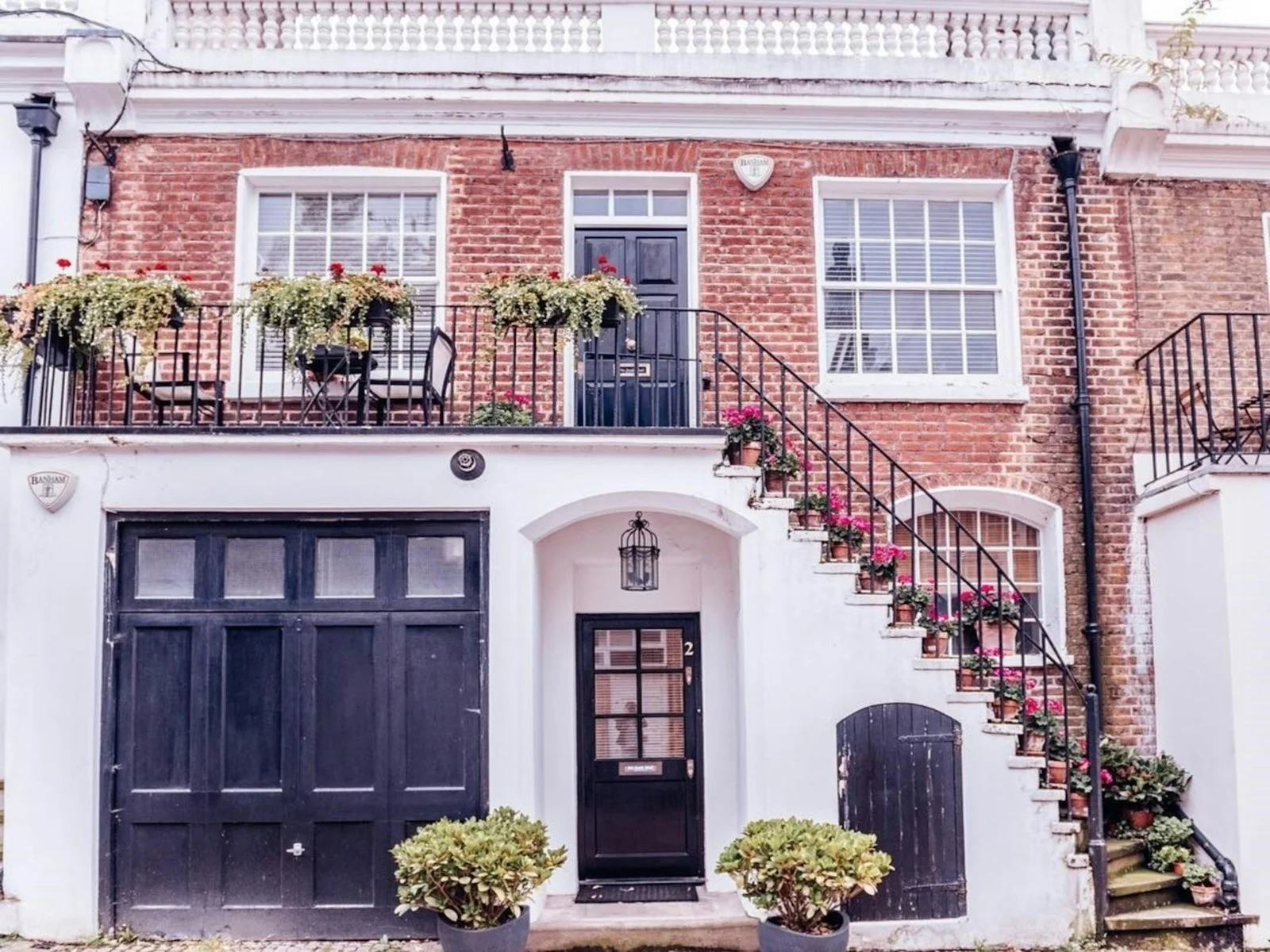 Red brick mews house with black garage and front doors, potted geraniums and burglar alarm boxes