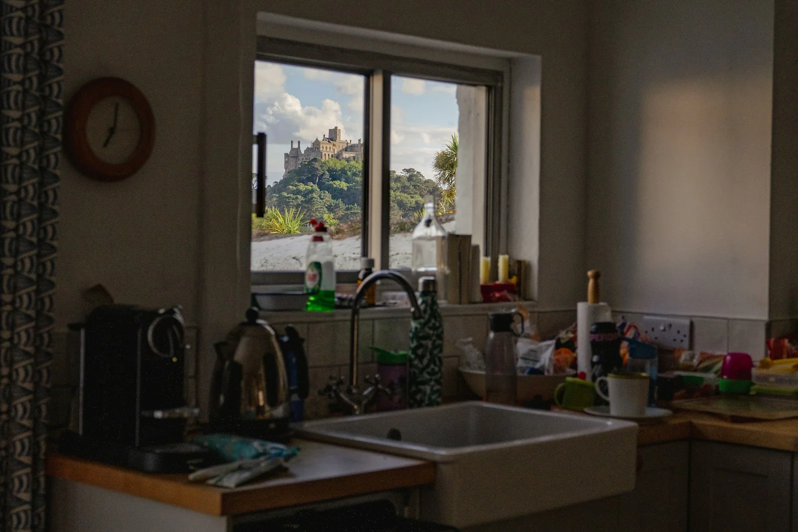 Cluttered kitchen with white butler sink and wooden worktops, window overlooking a hilltop castle